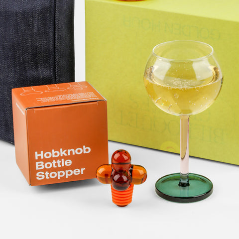 Close up of a wine glass with a pink stem and green bottom, an orange bottle stopper and a denim wine tote