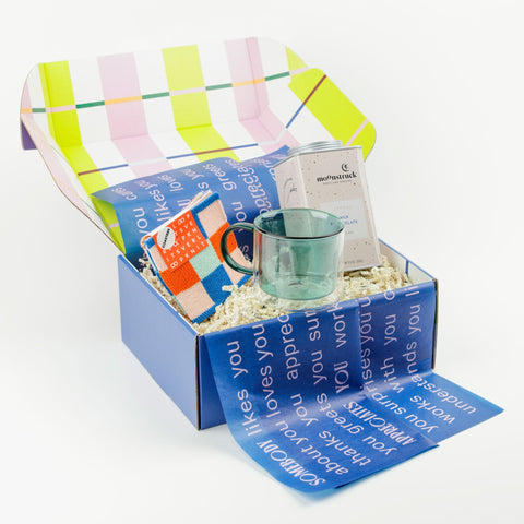 Teal glass mug with hot chocolate tin and colorful coasters in a gift box