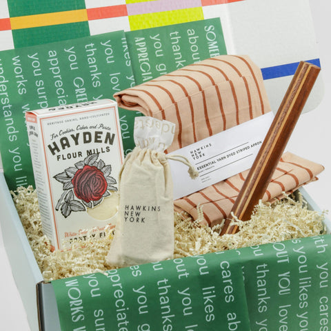 Awooden rolling pin, orange striped apron, a Hawkins New York linen bag and a box of pastry flour all in a colorful giftbox