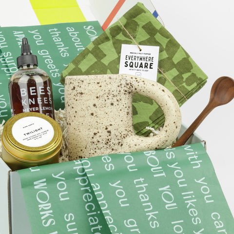 Light brown mug with dark brown speckles, tin candle, bottle of honey, green checked linen square and wooden coffee spoon in a box