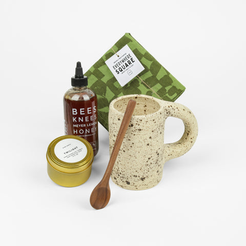 Wooden spoon, tin candle, bottle of honey, green checked linen square and a brown two toned speckled mug all sitting next to each other