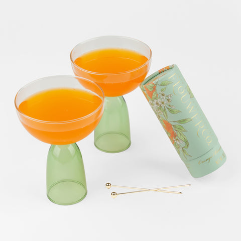 Two clear and green coupes with orange juice, two cocktail picks and a green tube of cocktail tubes leaning up against the coupe