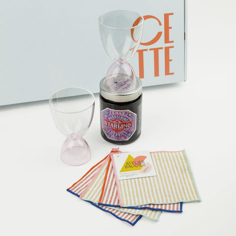 Two clear and pink glasses with a jar of cherries and striped taupe and red linen napkins with colorful edging in front of a CETTE box