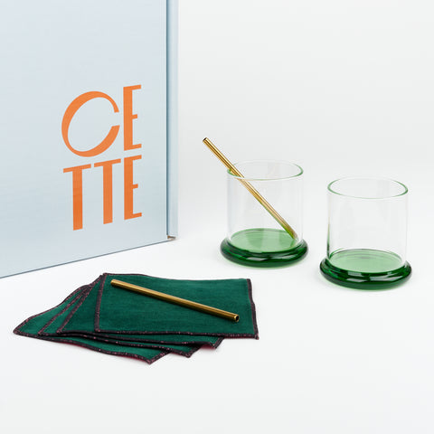 a pair of cocktail glasses with rounded green glass bases with two metal cocktail straws and a set of green velvet cotton cocktail napkins next to a pale blue Cette gift box
