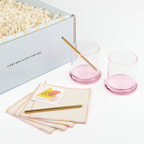 two sophie lou jacobsen cocktail glasses with rounded pink glass bases with two metal cocktail straws and a set of pink cotton cocktail napkins by Atelier Saucier next to an open gift box