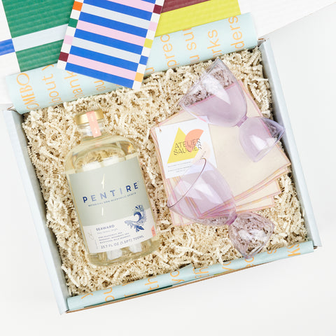 an open gift box featuring Atelier Saucier pink cocktail napkins, a pair of lavender Mamo classic glasses and a bottle of Pentire Seaward non-alcoholic aperitif