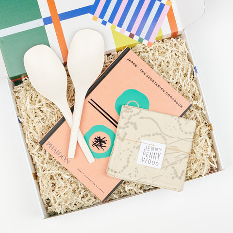 a gift box showcasing two white bamboo serving spoons, a Japanese vegetarian cookbook and a cotton linen tea towel with a natural white and fog colored abstract pattern
