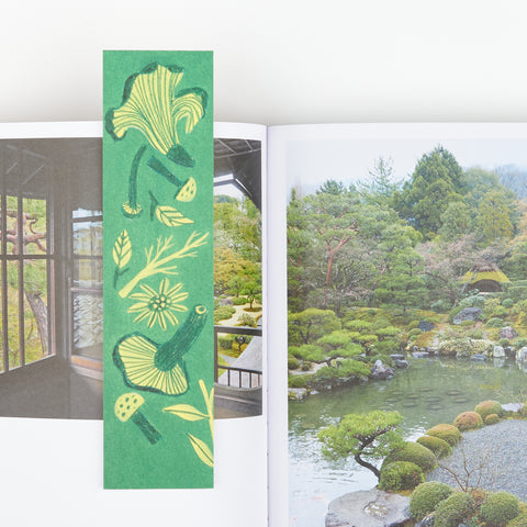 An bookmark with hand drawn nature and mushroom designs sitting on a open book showing two Japanese gardens