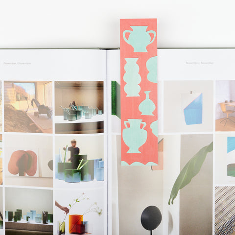 An illustrated red bookmark with cut outs of pale green vases sitting on an open design book with a variety of images