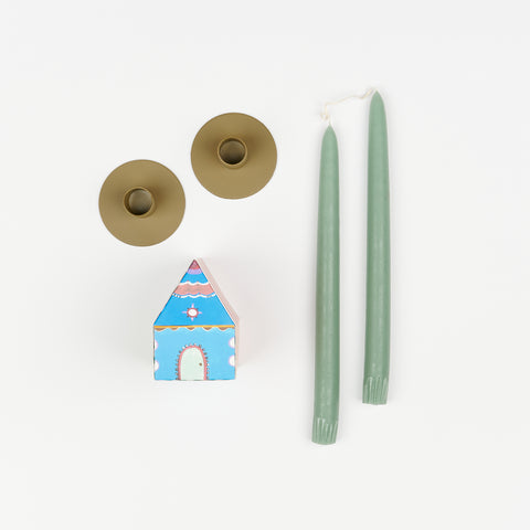 Sage green taper candles, pair of off-olive metal Hawkins New York Candle Holders, and State the Label box of matches in the shape of a house with colorful illustrations