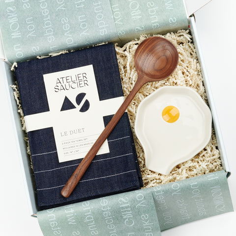 An open gift box featuring a gift set with a set of denim tea towels, a walnut spoon and a ceramic fried egg-like spoon rest