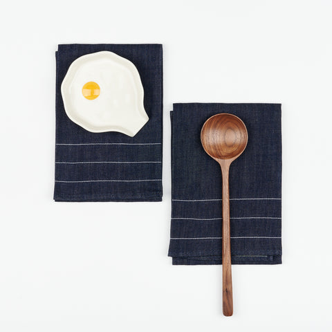 Two Atelier Saucier denim tea towels side by side, one has a Reva Preven Egg spoon rest on it, the other has a wooden round head walnut cooking spoon on it