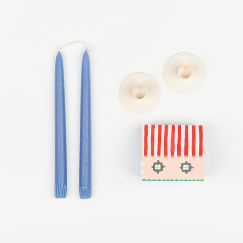 Light blue taper candles, pair of off-white metal Hawkins New York Candle Holders, and State the Label box of matches in the shape of a house with colorful illustrations