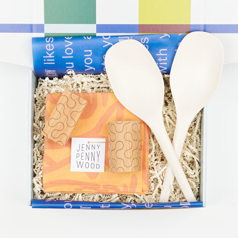 a gift box open to reveal two white Ekobo bamboo serving spoons, an orange and pink tea towel with an abstract design and a part of wooden salt and pepper shakers