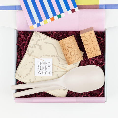 a gift box with plum colored crinkle paper and lilac tissue showing two stone gray serving spoons, a pair of wooden salt and pepper shakers and a white and light gray cotton linen Jenny Pennywood tea towel