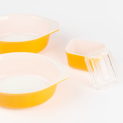 close up of two larger vintage Pyrex serving bowls and one smaller pyrex container with a glass lid leaning on its side. all are bright cheerful yellow on the outside and white on the interior