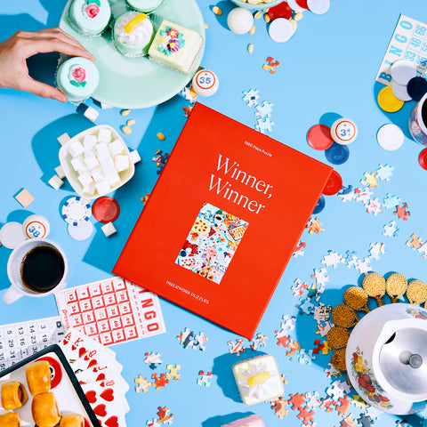 Red Winner, Winner Piecework Puzzle 1000 piece puzzle box surrounded by puzzle pieces, finger foods, bingo chips, playing cards, and desserts