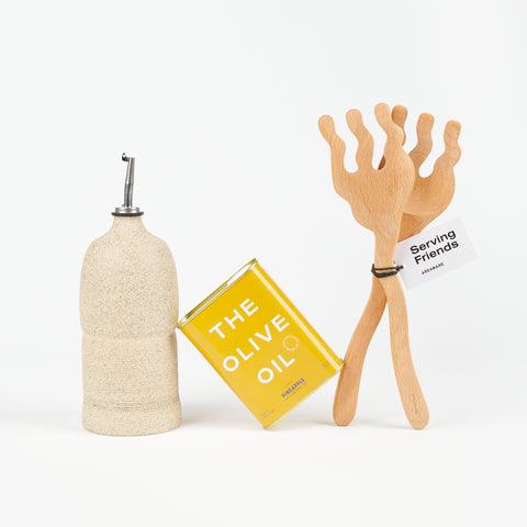 a golden yellow tin of Olive oil that is between an off-white ceramic oil cruet and a pair of wooden salad tongs that look like they have fingers on the end of them
