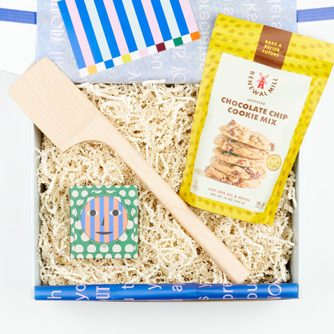 an open gift box with white crinkle filling and blue tissue paper featuring a kitchen timer in a box, a maple wood spatula and a bag of chocolate chip cookie mix