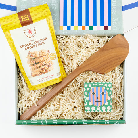 an open gift box featuring a bag of chocolate chip cookie mix, a walnut wood spatula, and a kitchen timer in a box with a colorful smiling geometric face