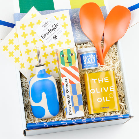 A Pantry gift box open and featuring two sweedish dish cloths with yellow crosses, two orange serving spoons, an oil cruet with a blue line design, an pepper grinder, a jar of flake salt and a yellow tin of olive oil