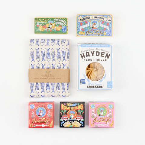 An arrangement of colorful Fishwife Tinned Fish boxes including Smoked Salmon, Spicy Salmon, Smoked Albacore Tuna, Anchovies, and Sardines above and below a cotton hand towel with blue fish and a box of White Sonora Crackers