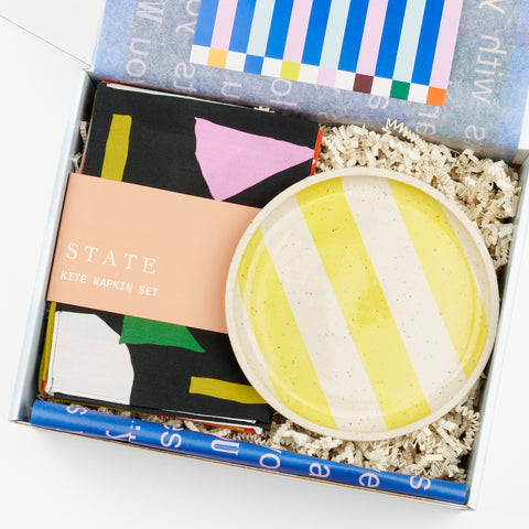 An open gift box with a set of State the Label Kite Napkins and a yellow striped ceramic plate