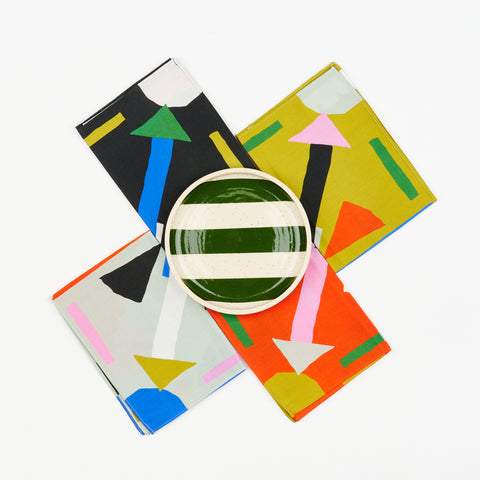 A set of four colorful napkins by State the Label laid out in an X shape and in the center of the X there is a ceramic plate with dark green stripes