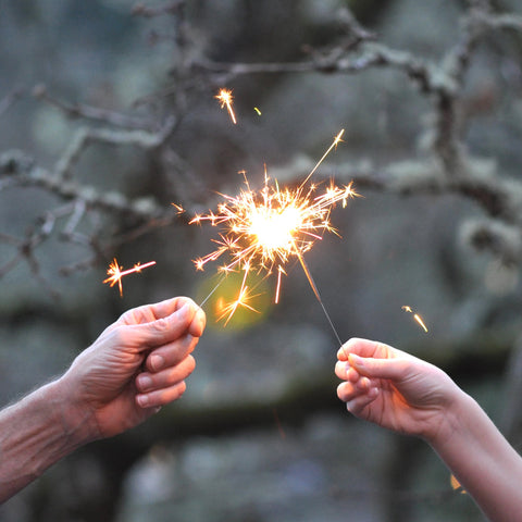 two hands holding metal sparklers that are lit and show sparks flying from the meeting of the two sparklers