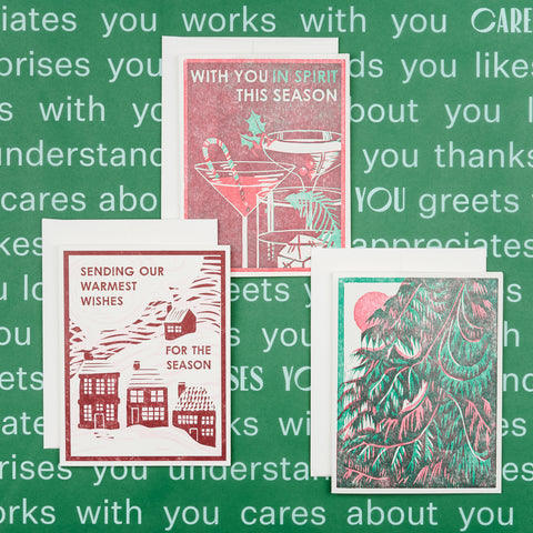 A Heartell holiday card trio, one Sending Our Warmest For The Season card, one of a green fir tree and another with a red background, drinks and With You In Spirit This Season