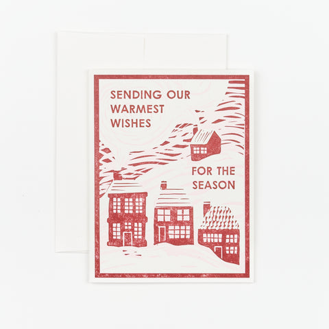 A blank holiday notecard with an illustration of red houses in snow that reads "Sending our warmest wishes for the season" on the front of the card