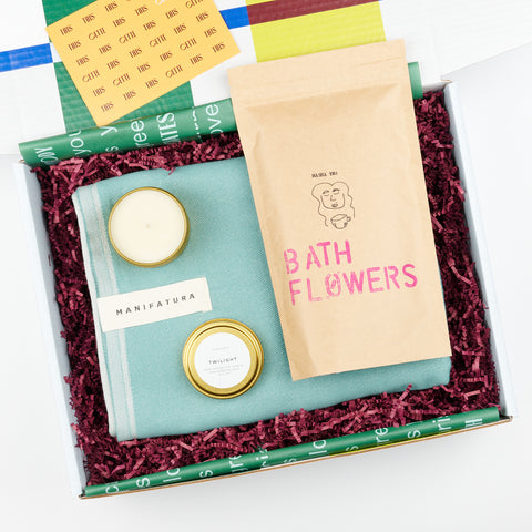 open gift box with green bath towel, open tin candle and paper pouch of bath flowers