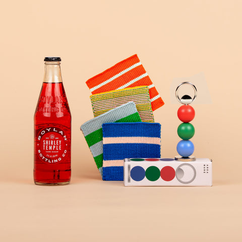 Front view of Pop of Color gift set showing a set of Verloop striped knit coasters, a colorful bottle opener by Areaware, and a bottle of Boylan’s Shirley Temple soda on a neutral background.