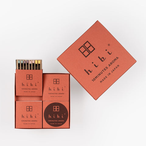 Overhead view of an open Hibi incense gift box, revealing assorted fragrance boxes with black incense matches topped with rainbow-colored match heads and a wool burning pad.