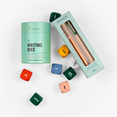 Overhead flat lay of The Writer’s Block Gift Set showing Papier ballpoint pens in packaging and Two Tumbleweeds Writing Dice partially rolled out from their container.