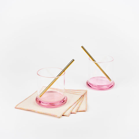 A pair of Sophie Lou Jacobsen fritter glasses with pink bases, two metal cocktail straws and a set of four pink hummingbird Atelier Saucier cocktail napkins
