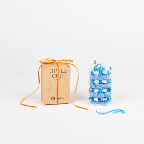 A Sophie Lou Jacobsen clear Ripple cup filled with blue crinkle paper next to a brown ripple cup box with a thin orange ribbon around it