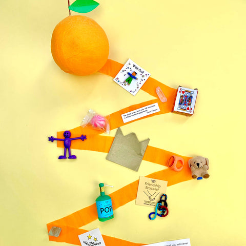 TOPS Malibu orange surprise ball partially unwound showing mini treats like playing cards, fun toys, and a paper crown on a yellow background