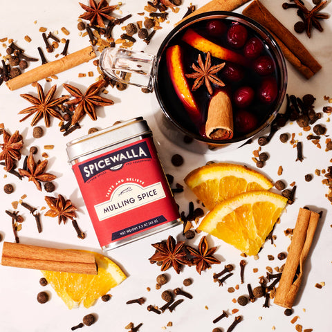Spicewalla mulled spice mix tin with a mug of mulled wine, oranges, and spices on a white background