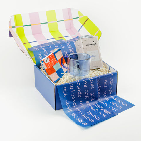 Gift box that has a blue glass mug, a tin of hot chocolate and colorful coasters