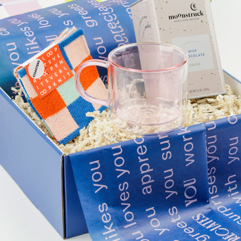 Close up view of a pink glass mug, a tin of hot chocolate and colorful coasters in a gift box