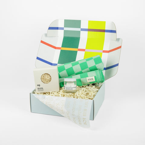 A green checkered towel, a glass jar of shower steams and a body sponge in a colorful box