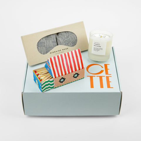 A dark blue eye mask, candle and colorful match house all on top of a box