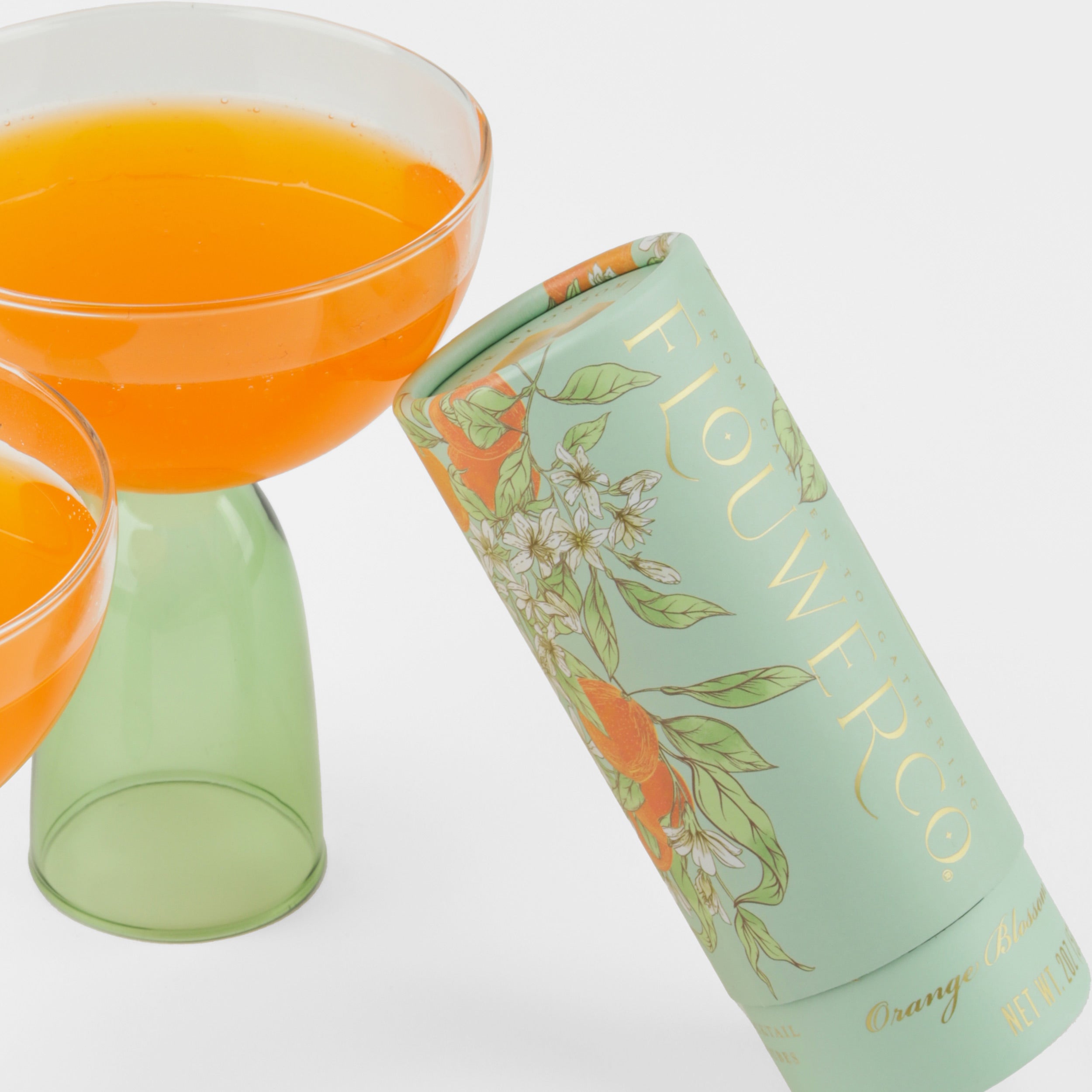 Close up of a clear and green coupe with orange juice and a green tube of cocktail cubes