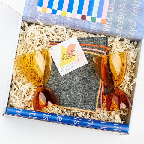 a gift box showing a set of four Atelier Saucier chambray cloth cocktail napkins and two honey and amber Mamo classic glasses