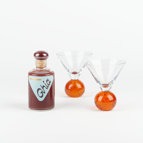 A bottle of Ghia Aperitif next to two vintage short martini glasses with round bright orange glass bases