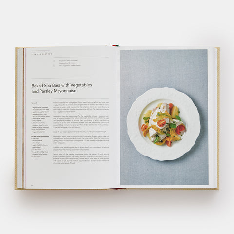 Eataly Italian Cookbook open and showing a recipe for Baked Sea Bass and Vegetables and a photograph of the meal on a white plate sitting on a blue table cloth