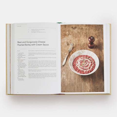 Eataly Italian Cookbook open showing a recipe for Beet and Gorgonzola Pearled Barley dish with a photograph of the dish in a white bowl on a wooden table with a fork and pepper grinder