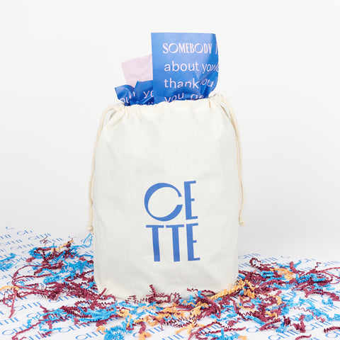 a white drawstring bag that reads CETTE in blue letters with blue and lilac tissue paper sticking out of it surrounded by multi-colored crinkle paper