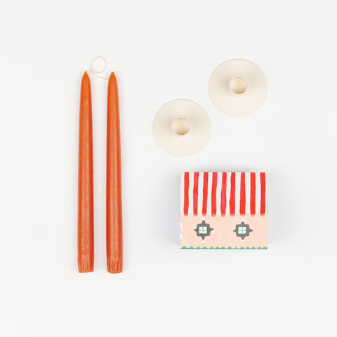 Burnt orange taper candles, pair of off-white metal Hawkins New York Candle Holders, and State the Label box of matches in the shape of a house with colorful illustrations